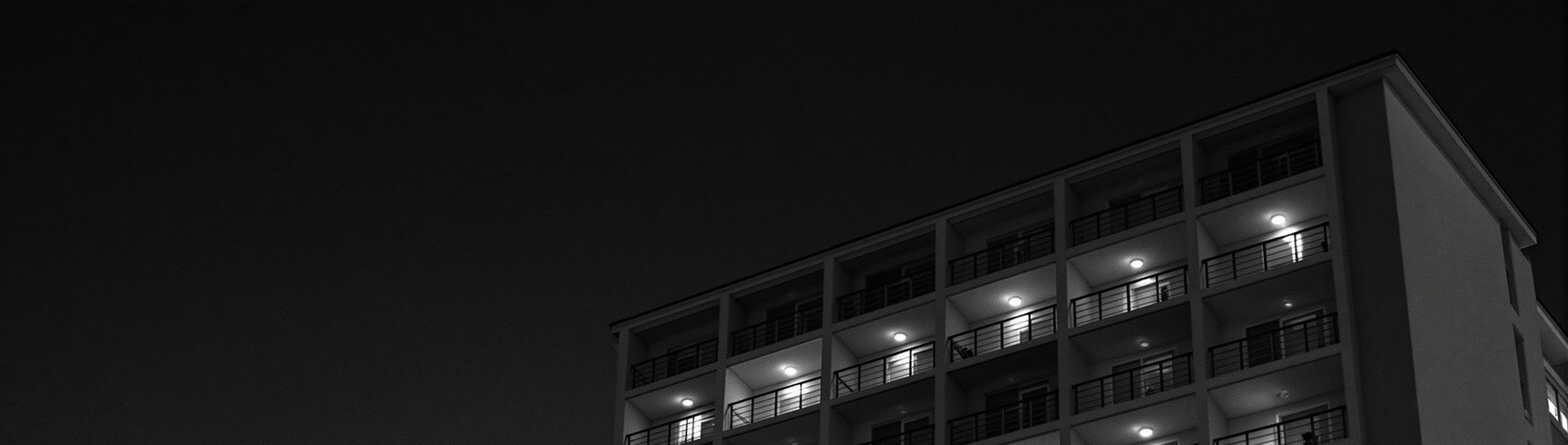 Fachada de un hotel modernista de los setenta al anochecer, con la luz cálida
        saliendo de las habitaciones por los balcones.